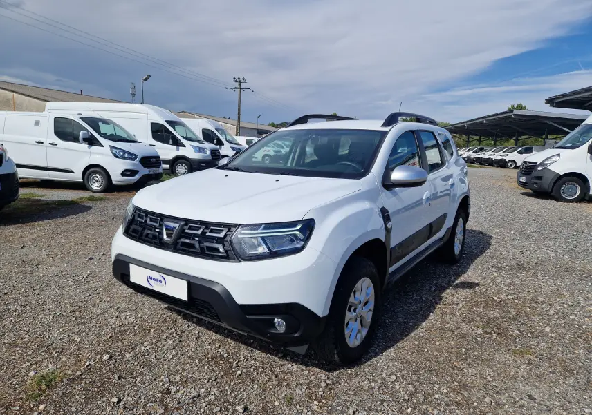 Dacia Duster blanc vue 3/4 avant droit, SUV compact avec protections noires et barres de toit visibles.