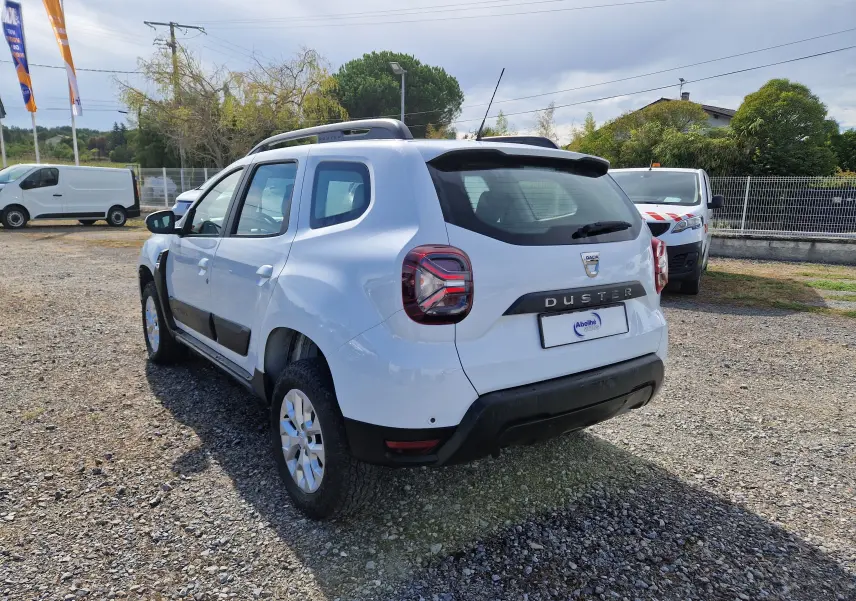 Vue 3/4 arrière droite du Dacia Duster blanc 2023 avec barres de toit et protections noires sur les portes.