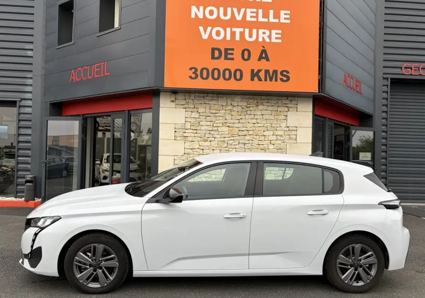 Profil droit d'une Peugeot 308 blanche 2024 garée devant un bâtiment avec enseigne orange "Votre nouvelle voiture de 0 à 30000 kms".