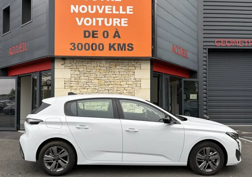 Profil latéral droit d'une Peugeot 308 blanche 2024 garée devant un bâtiment avec une grande enseigne orange.