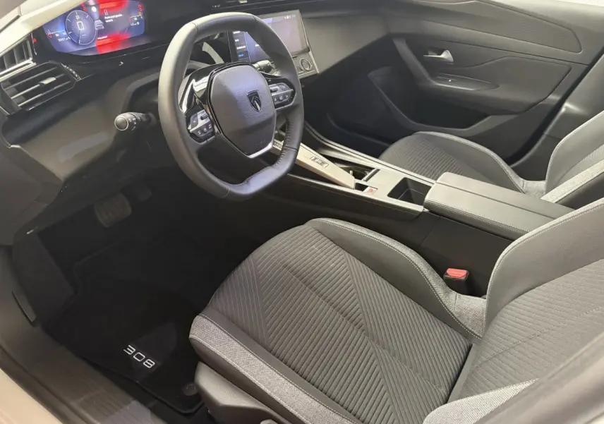 Intérieur de la Peugeot 308 2024 vu côté conducteur, sièges gris, volant cuir multifonctions et tableau de bord digital.