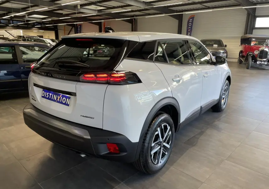 Peugeot 2008 hybride blanche vue 3/4 arrière droit en intérieur, feux arrière LED allumés et jantes bicolores.