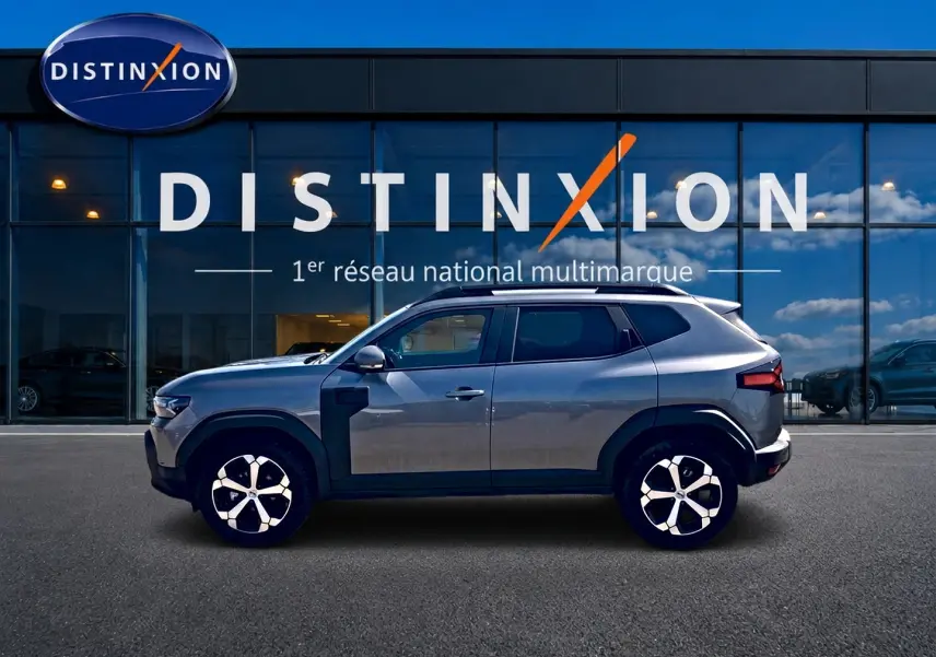 Profil droit du Dacia Duster gris schiste 2026 avec jantes alu et barres de toit, devant une vitrine Distinxion.