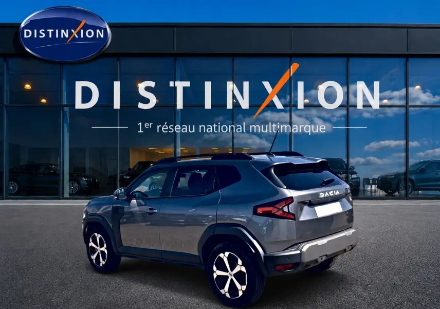 Vue 3/4 arrière droite du Dacia Duster gris schiste 2026 devant une vitrine Distinxion, avec jantes alu et barres de toit.