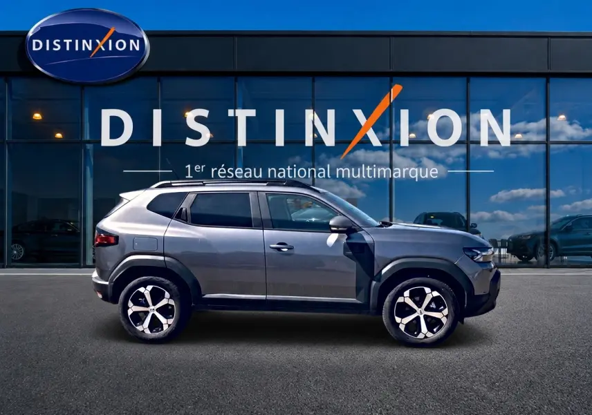 Profil droit du Dacia Duster gris schiste 2026 avec jantes alu et barres de toit devant vitrine Distinxion.