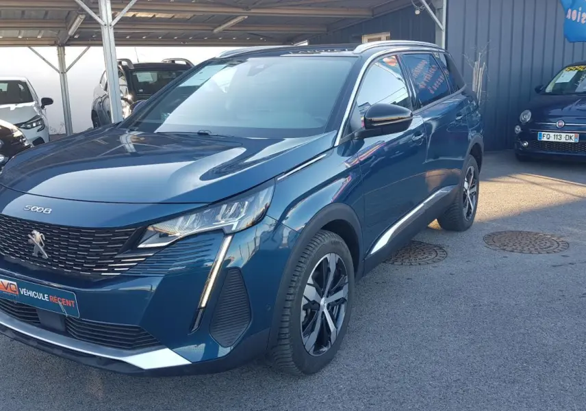 Peugeot 5008 bleu en 3/4 avant droit, avec jantes alliage 18 pouces et barres de toit visibles.