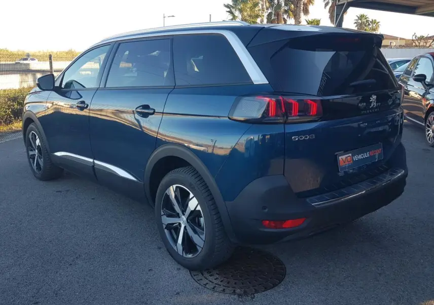 Peugeot 5008 bleu en 3/4 arrière droit, avec jantes alliage 18 pouces et barres de toit visibles.