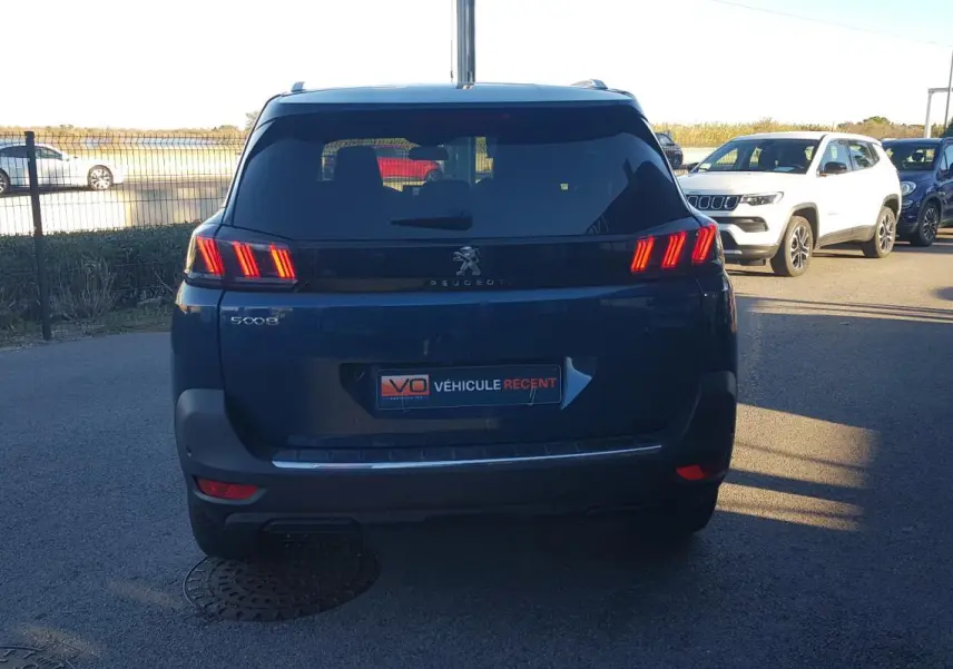 Vue arrière d’un Peugeot 5008 bleu 2024 avec feux allumés et barres de toit visibles en extérieur.