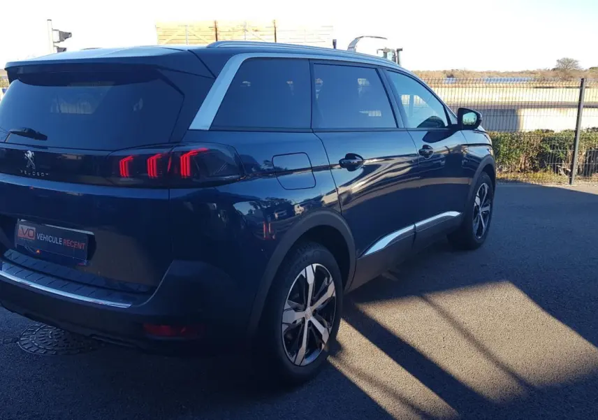 Vue 3/4 arrière droite d’un Peugeot 5008 bleu 2024 avec jantes alliage 18 pouces et barres de toit visibles.