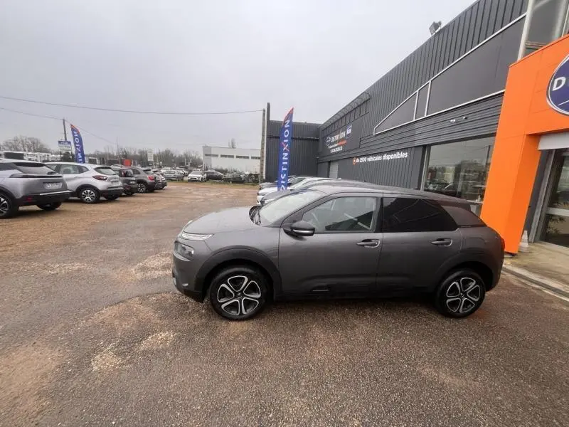 Vue de profil côté gauche d'une Citroën C4 Cactus gris Platinium avec jantes spécifiques et vitres teintées, stationnée devant un bâtiment.