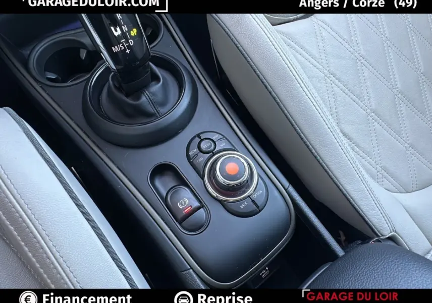 Gros plan sur la console centrale noire avec molette de commande et levier de vitesse, sièges cuir gris clair visibles.