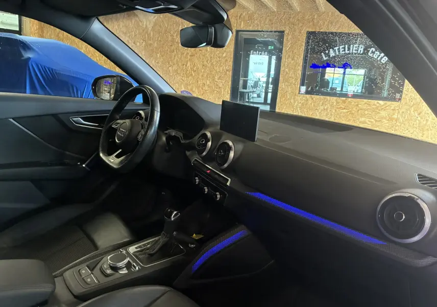 Intérieur noir de l'Audi Q2 2023, vue côté conducteur, avec éclairage d'ambiance bleu et tableau de bord moderne.