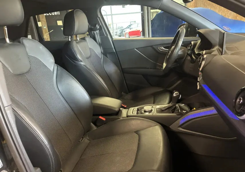 Vue intérieure côté conducteur de l'Audi Q2 S-Line noire, sièges sport cuir noir et éclairage d'ambiance bleu sur tableau de bord.