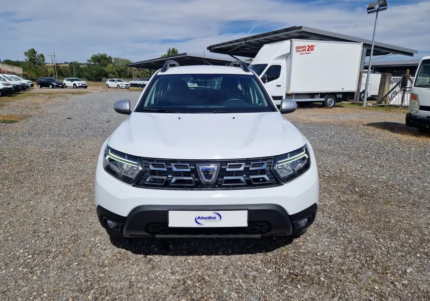 Vue avant d'un Dacia Duster 4x4 blanc 2023 avec calandre noire et phares LED allumés sur un sol gravillonné.