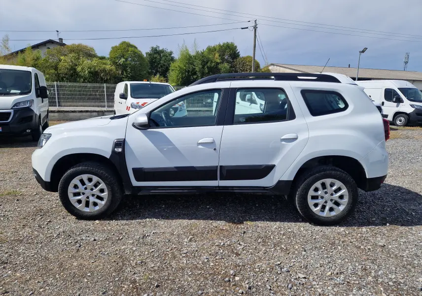 Profil côté gauche d'un Dacia Duster blanc 4x4 2023, avec protections noires sur les portières et jantes alliage 16 pouces.