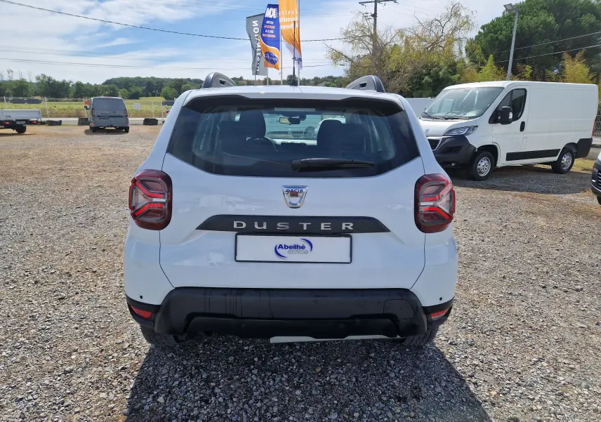 Vue arrière du Dacia Duster blanc 2023 avec logo et feux arrière distinctifs sur un terrain gravillonné.