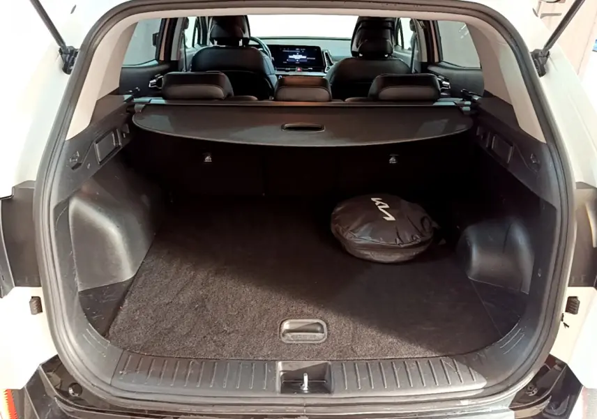 Coffre ouvert d'un KIA SPORTAGE blanc vu de l'arrière, avec cache-bagages et sac noir marqué KIA visible à l'intérieur.