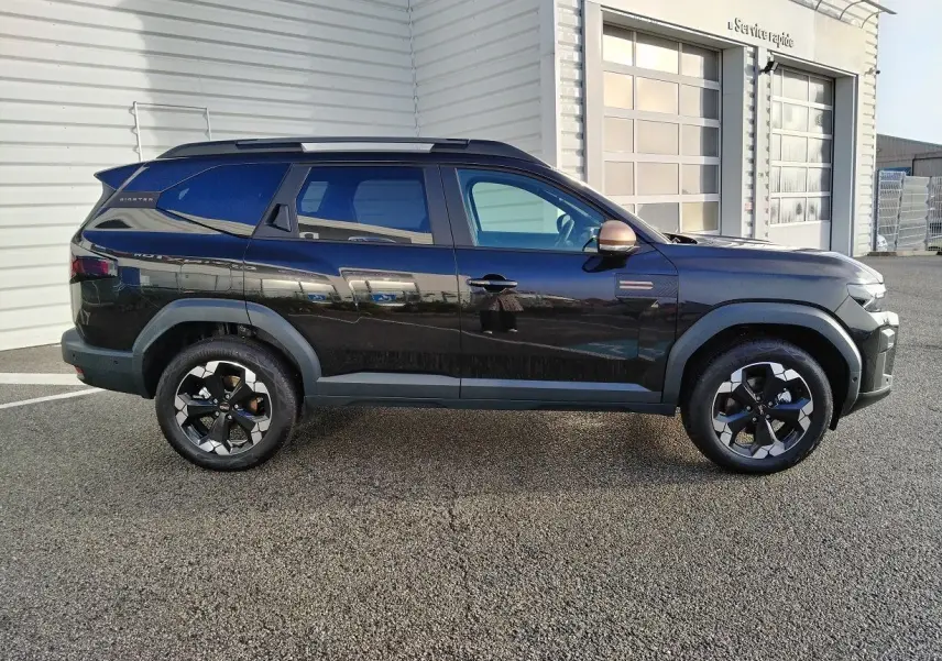 Profil droit d'un Dacia Bigster noir 2025 avec jantes bicolores et rétroviseurs cuivrés, stationné devant un bâtiment blanc.