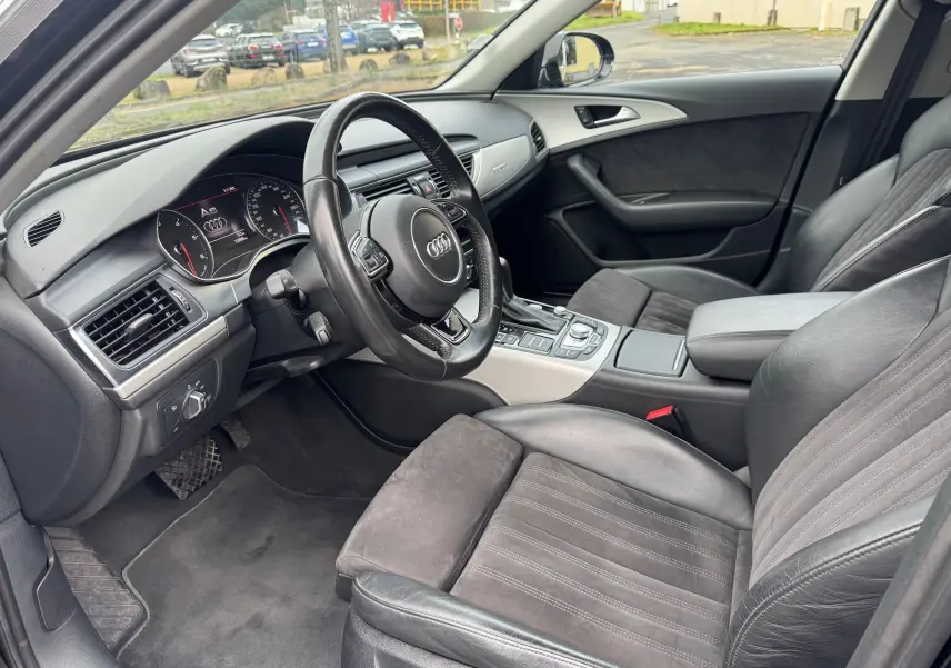Vue intérieure côté conducteur de l'Audi A6 Avant 2015 noire, avec sièges cuir et alcantara, volant multifonction et tableau de bord digital.