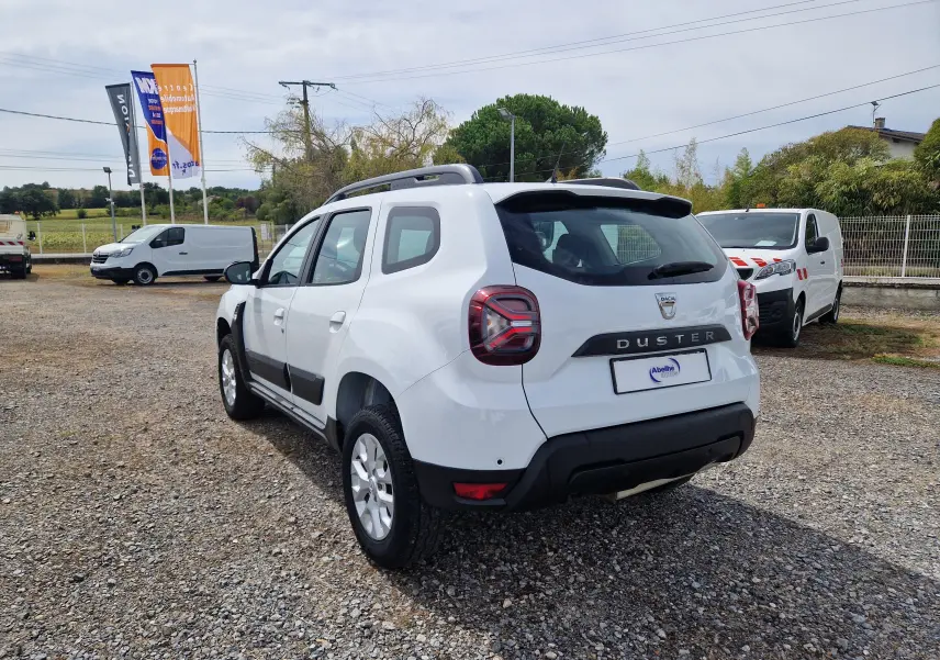 Vue 3/4 arrière droite d'un Dacia Duster blanc 4x4 2022 avec barres de toit et protections noires sur un parking.