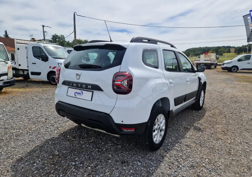 Vue 3/4 arrière droite d'un Dacia Duster blanc 4x4 avec barres de toit et protections latérales noires.