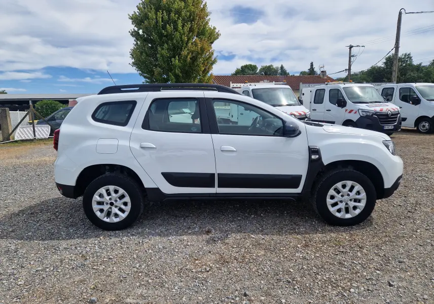 Profil côté gauche d'un Dacia Duster blanc 4x4 avec barres de toit noires, sur un sol gravillonné en extérieur.