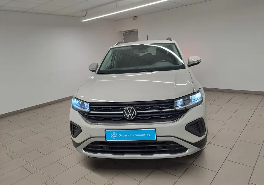 Vue avant d'un Volkswagen T-Cross 2024 gris Ascot avec phares allumés dans un showroom blanc.