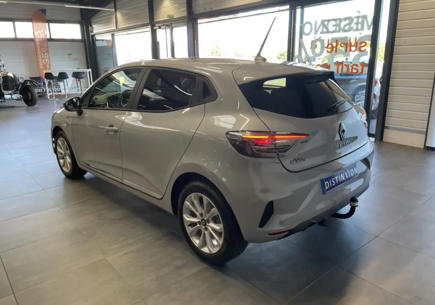 Renault Clio 1.5 Blue dCi 2025 gris Rafale vue 3/4 arrière droit en intérieur avec feux arrière allumés.
