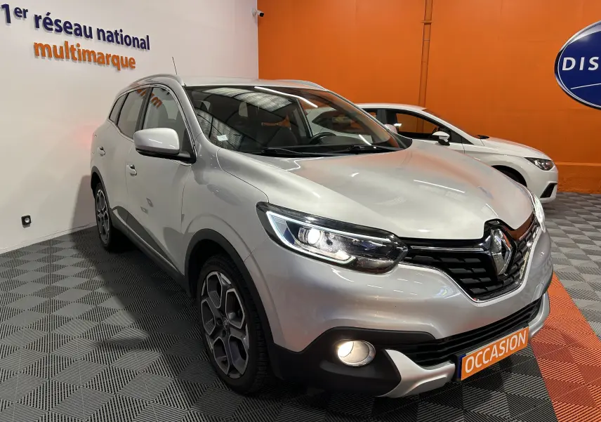 Vue 3/4 avant d'un Renault Kadjar gris avec phares allumés et plaque "OCCASION" en concession intérieure.
