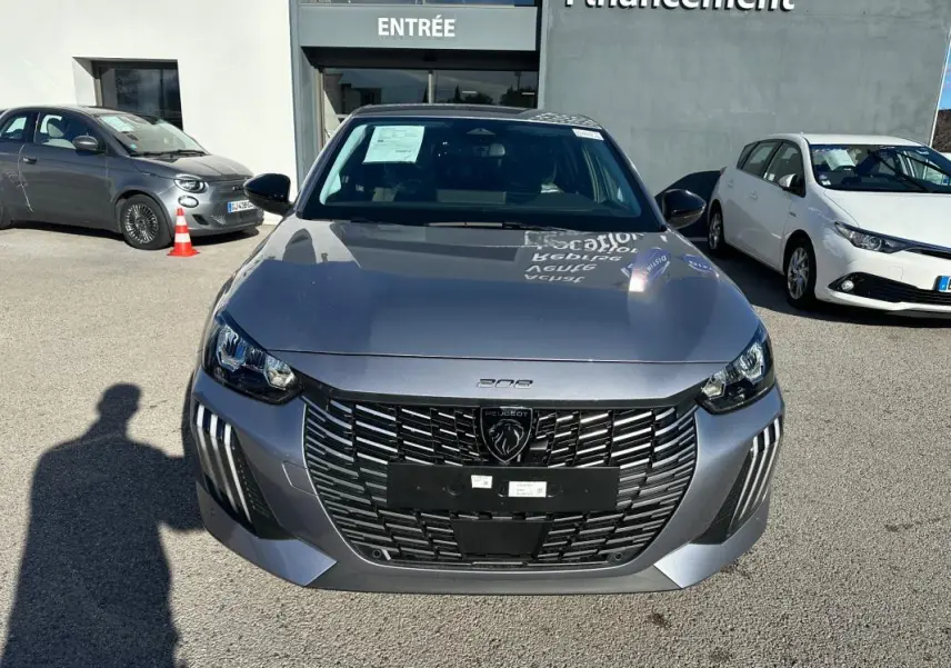 Vue avant d'une Peugeot 208 Hybrid 110 gris métallisé avec calandre noire et phares LED, stationnée devant un bâtiment.