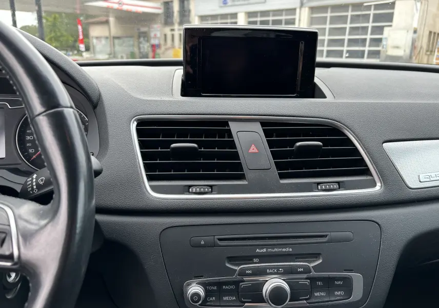 Vue intérieure centrée sur le tableau de bord noir de l'Audi Q3 2014 avec écran multimédia et commandes centrales visibles.