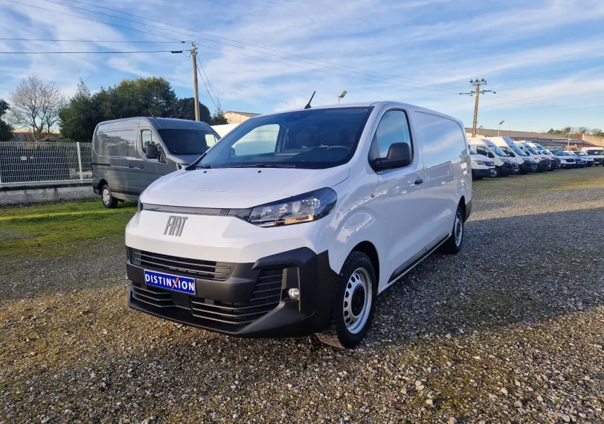 Fourgon FIAT Scudo blanc en 3/4 avant droit, avec pare-chocs noirs et jantes acier, stationné sur un terrain gravillonné.