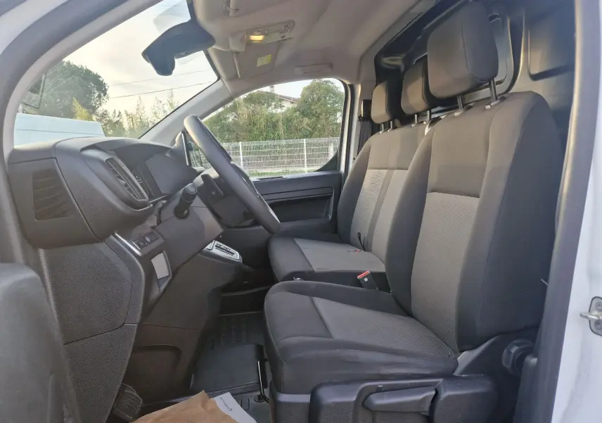 Vue intérieure côté conducteur du Fiat Scudo Fourgon blanc 2024, montrant les sièges avant en tissu gris et noir et le tableau de bord.