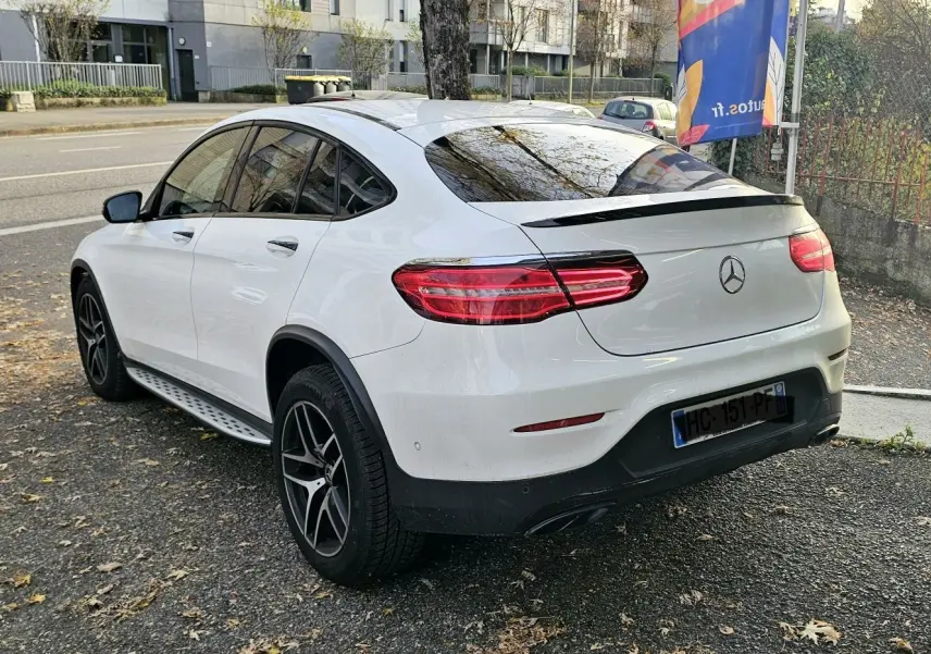 Vue 3/4 arrière droite d'un Mercedes GLC Coupé blanc avec jantes noires et vitres teintées sur parking urbain.