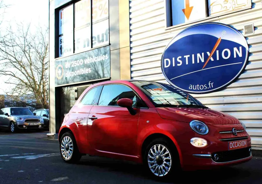 Vue 3/4 avant droite d'une FIAT 500C 2018 rouge pastel avec phares allumés et plaque "OCCASION" devant un concessionnaire.