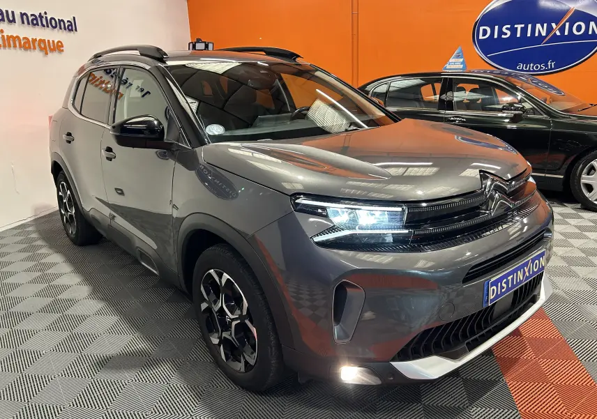 Citroën C5 Aircross gris Platinium en 3/4 avant droit, phares allumés, dans un showroom avec fond orange.