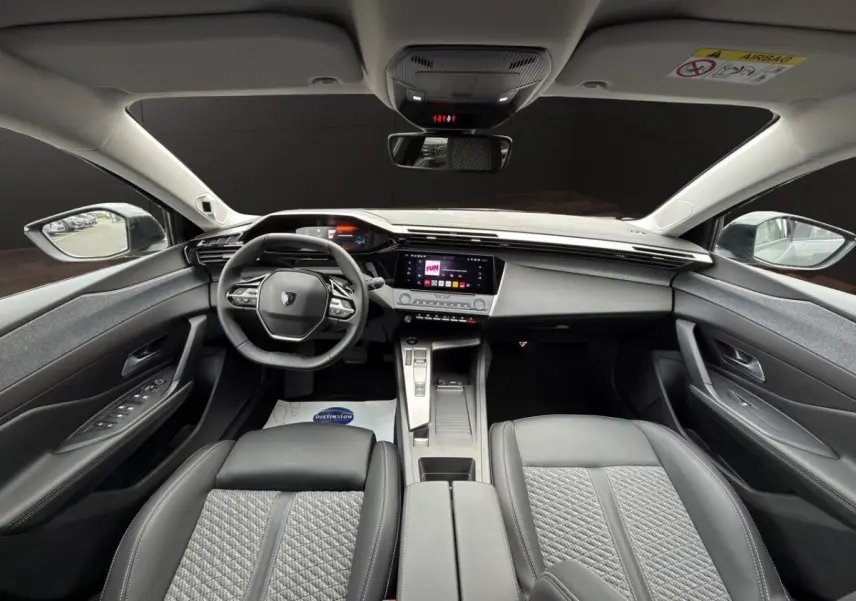 Vue intérieure avant de la Peugeot 308 Hybrid 145 Allure 2025, avec tableau de bord moderne et sièges gris foncé en tissu et cuir.
