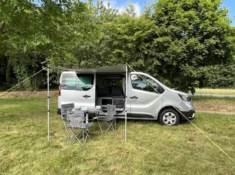 Vue latérale droite d’un Renault Trafic Fourgon gris 2025 avec auvent déployé et mobilier de camping installé.