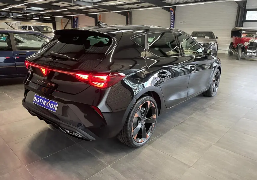 Vue 3/4 arrière droite d'une CUPRA Leon noire avec jantes cuivrées et feux arrière LED allumés en intérieur showroom.