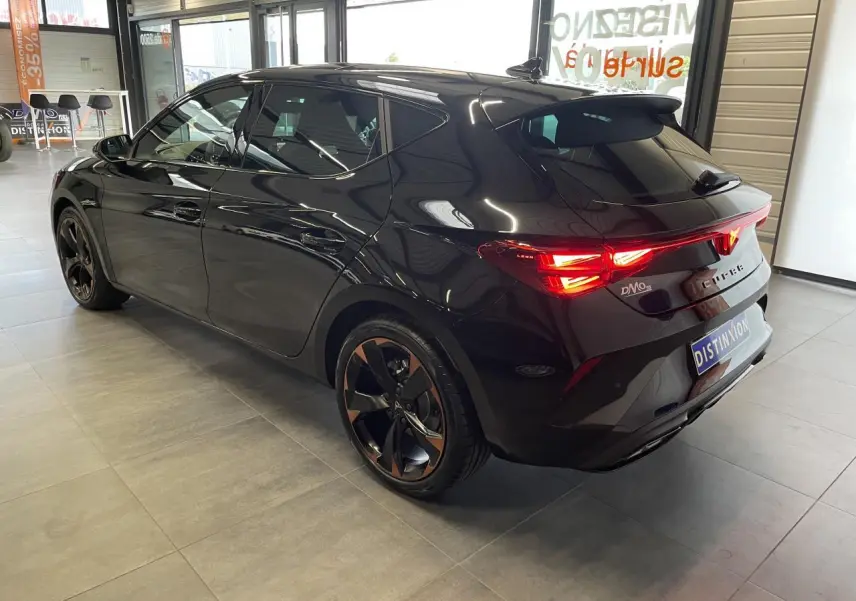 Vue 3/4 arrière droite d'une CUPRA Leon noire avec jantes cuivrées et feux arrière allumés en intérieur showroom.