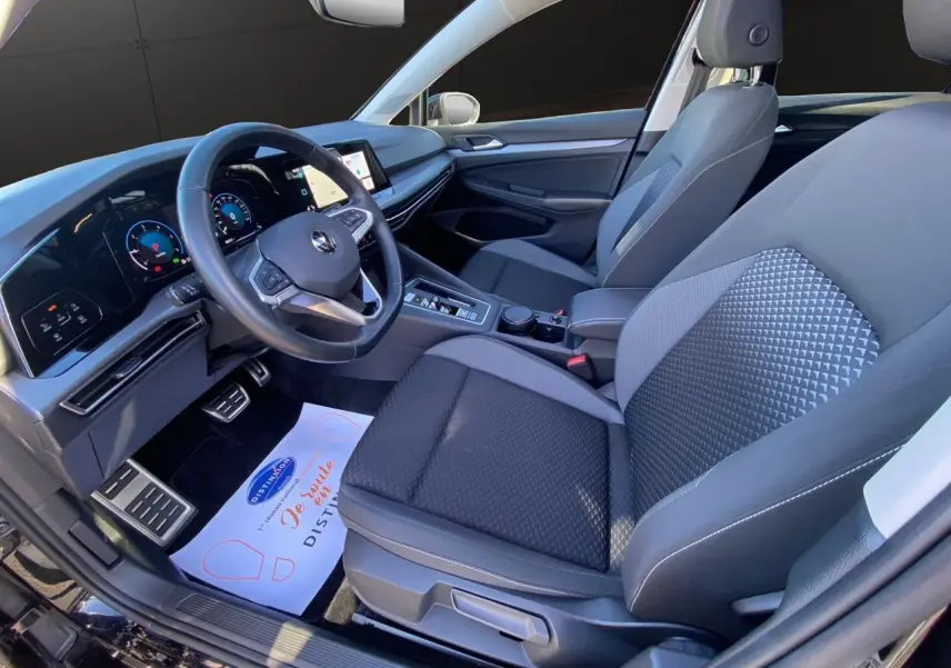 Vue intérieure côté conducteur de la Volkswagen Golf VIII noire, avec tableau de bord numérique et sièges tissu noir.