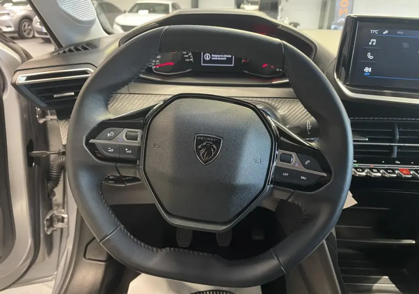 Vue intérieure centrée sur le volant cuir noir de la Peugeot 208 gris clair avec tableau de bord et écran tactile.