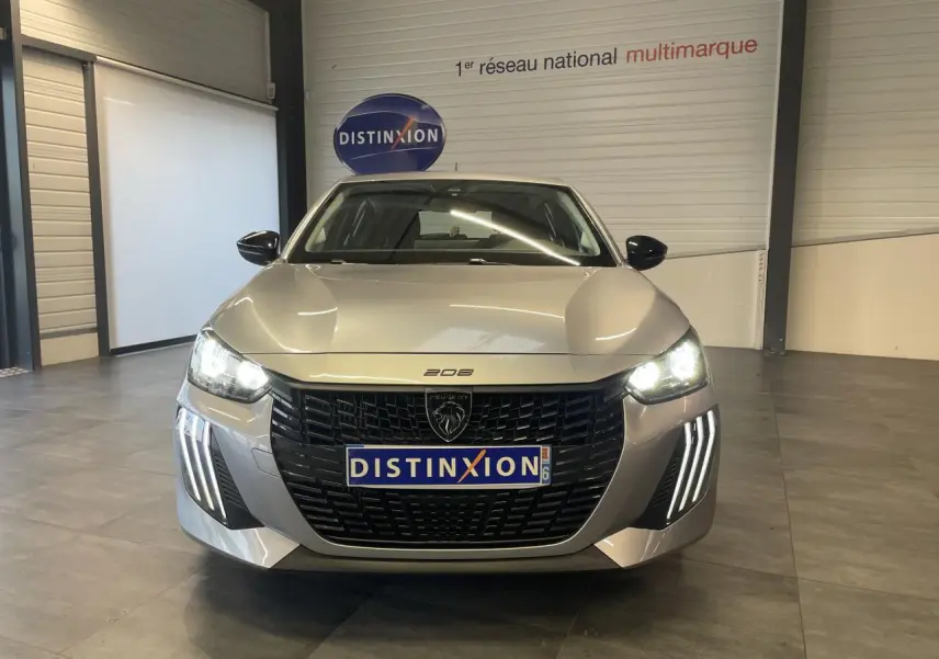 Vue frontale d'une Peugeot 208 gris clair 2024 avec calandre noire et feux LED allumés en intérieur showroom.