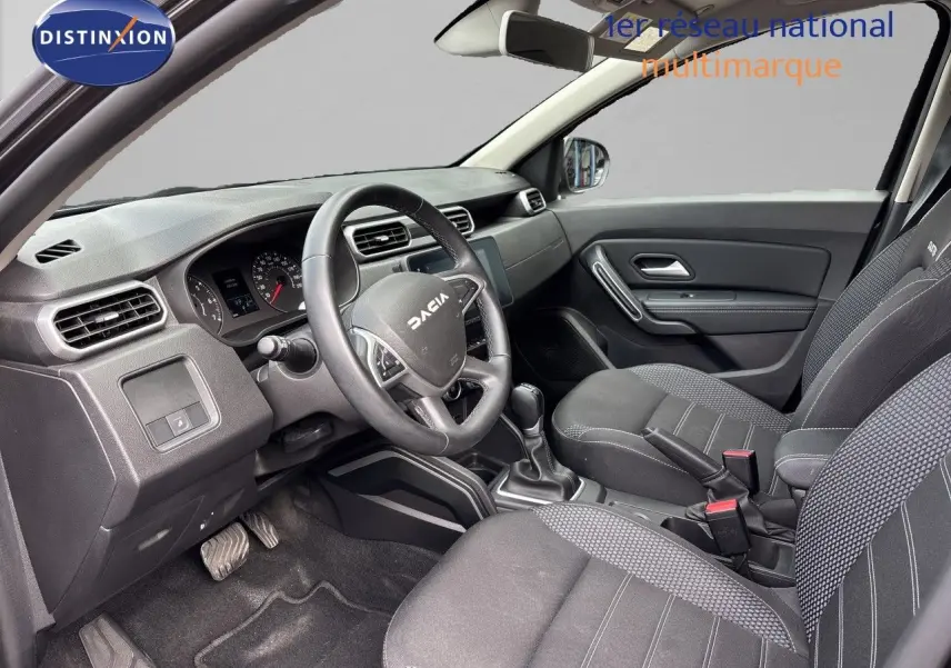 Vue intérieure côté conducteur du Dacia Duster 2024 avec tableau de bord noir et sièges tissu gris foncé.