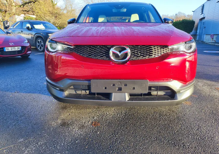 Vue frontale d'une Mazda MX-30 rouge métal 2023 avec calandre noire et phares LED allumés, stationnée en extérieur.