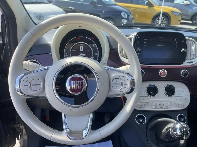 Vue intérieure centrée sur le volant beige et le tableau de bord de la Fiat 500 gris foncé, avec levier de vitesse manuel visible.