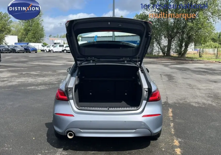Vue arrière d'une BMW Série 1 116i 2022 gris Frozen Pure avec coffre ouvert sur un parking extérieur.