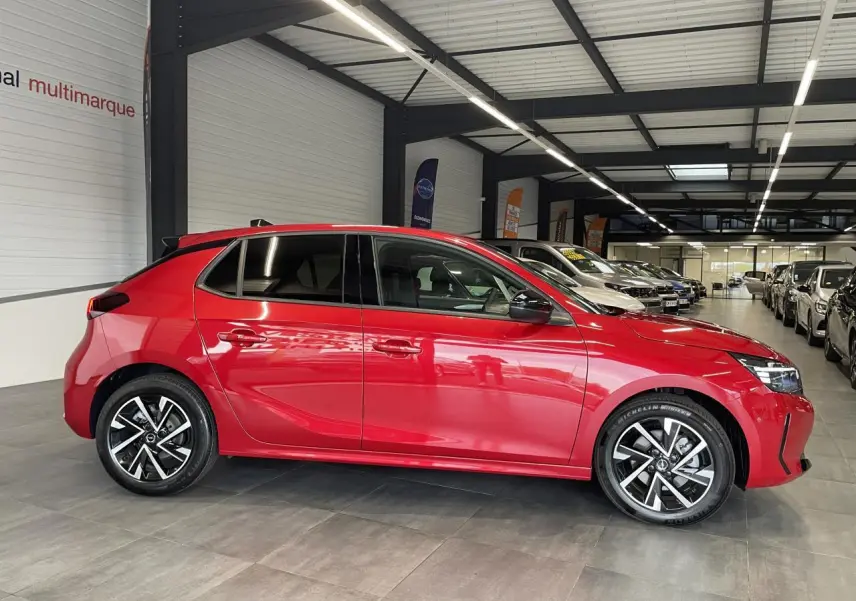 Vue de profil côté gauche d'une Opel Corsa rouge métallisée avec jantes alliage 16 pouces et vitres surteintées en intérieur showroom.