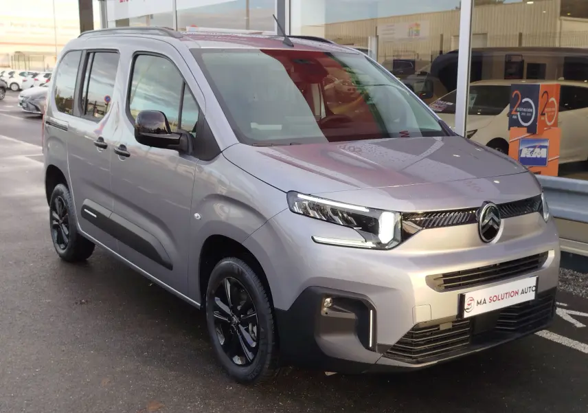 Vue 3/4 avant droit d'un Citroën Berlingo gris acier avec jantes noires et feux LED allumés, stationné devant un showroom.
