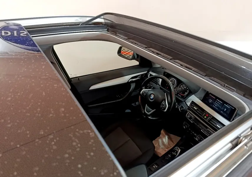 Vue intérieure du poste de conduite d'une BMW X1 noire 2020, avec volant cuir et écran tactile visible depuis le toit ouvrant.